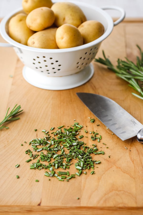 Fresh rosemary chopped on a cutting board and a white colander with yellow baby potatoes.
