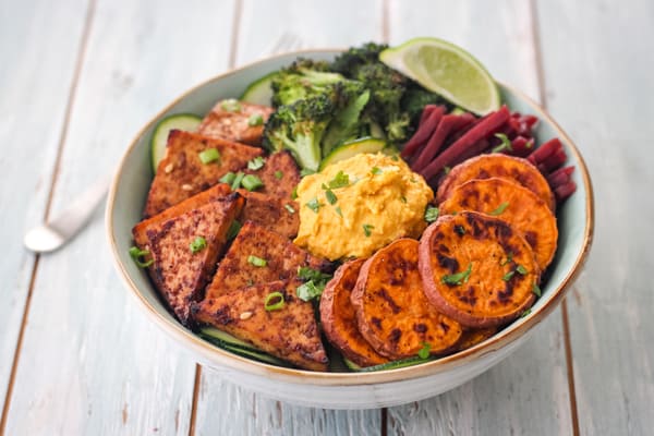 Broiled tofu in a large bowl filled with sweet potato rounds, broccoli and hummus.